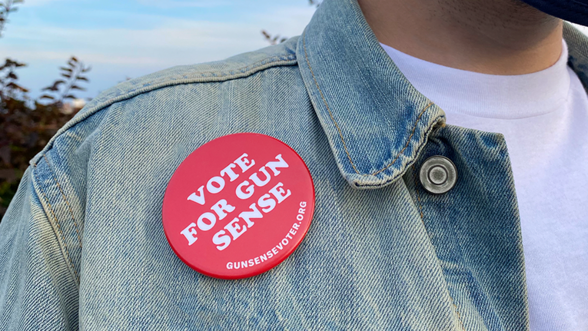 Featured image for the Gun Sense Voter Guide. A person wears a faded light-blue denim jacket and a white t-shirt. A red "Vote for Gun Sense" button is pinned on the jacket next to the right lapel. The button is fire-engine red and the text is white; the bottom of the button has "GunSenseVoter.org" printed on it.