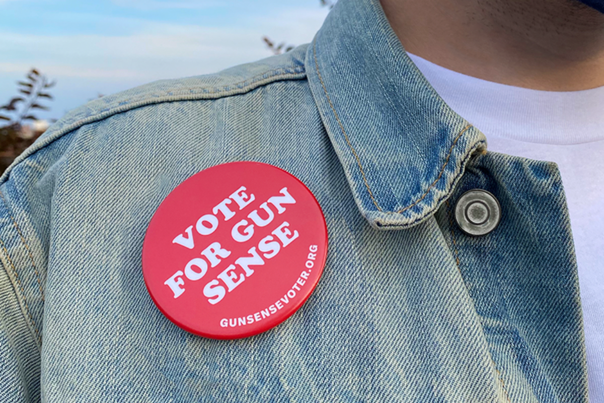 Featured image for the Gun Sense Voter Guide. A person wears a faded light-blue denim jacket and a white t-shirt. A red "Vote for Gun Sense" button is pinned on the jacket next to the right lapel. The button is fire-engine red and the text is white; the bottom of the button has "GunSenseVoter.org" printed on it.