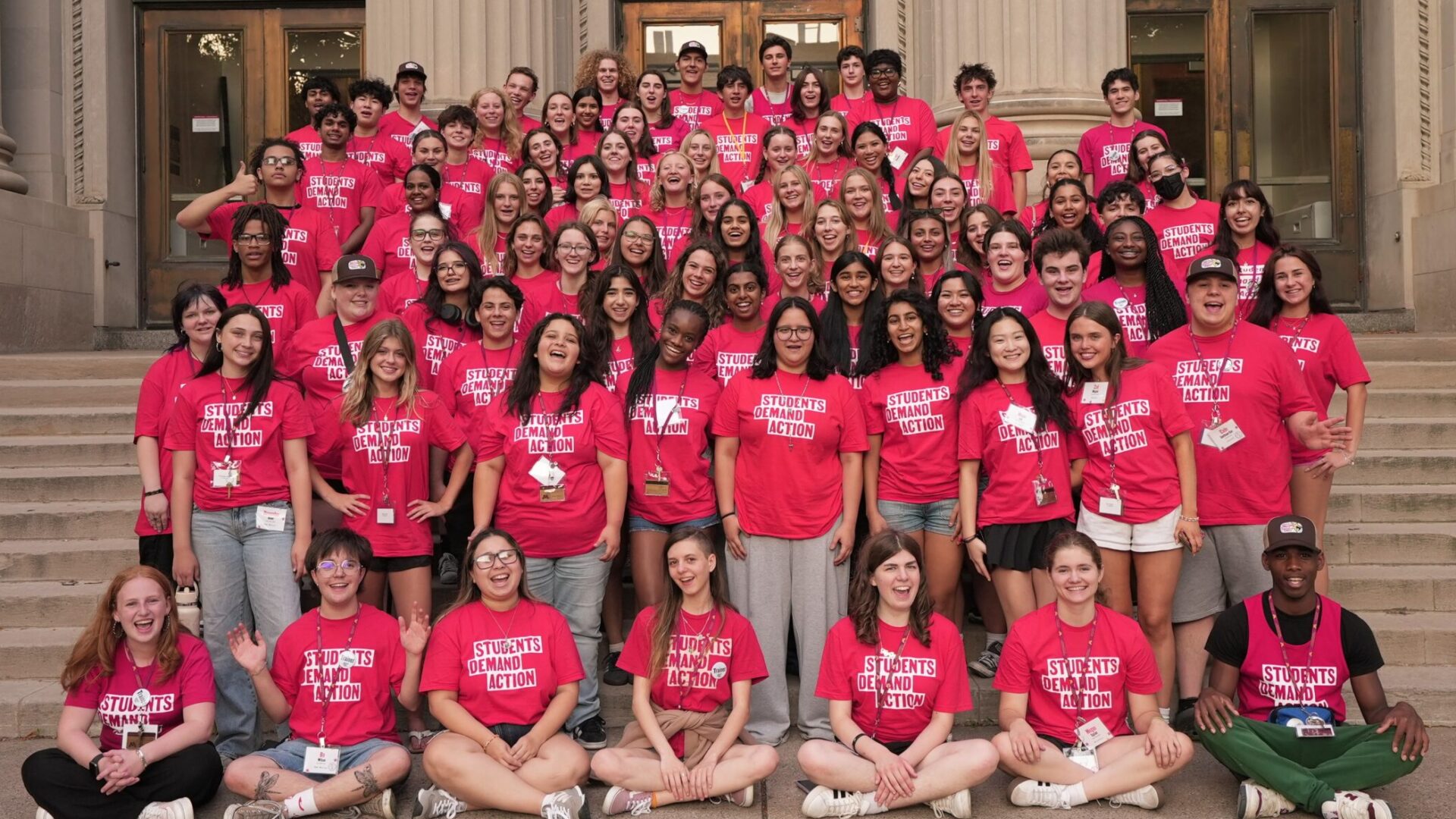 100 Students Demand Action volunteers in rows of 7 to 8 people pose for a group photo on the steps of a university campus building.