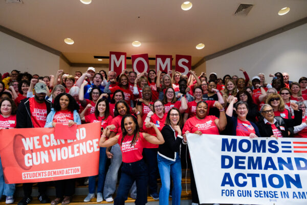A group of over 100 Moms Demand Action and Students Demand Action volunteers, all wearing red shirts, pose for a photo. Most are holding one arm up and flexing their bicep. Angela Ferrell-Zabala, executive director of Moms Demand Action and Students Demand Action, has her sleeves rolled up and is flexing in between two groups of volunteers who are holding "We Can End Gun Violence" Students Demand Action and "Moms Demand Action for Gun Sense in America" banners.